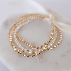 This stunning stack of dainty bracelets, feature a luxurious mix of gold-filled beads, freshwater pearls, and sparkly crystal zircon. This bracelet stack is a great set for instant impact or wear them separately to create multiple looks. This stack of five stretch bracelets includes: 1 - 4mm 14K gold filled bead bracelet 1 - 3mm 14K gold filled bead bracelet with silver floating heart 1 - 2.5mm 14K gold filled bead bracelet 1 - 3mm freshwater pearl stretch bracelet 1 - 3mm grade Aa  geuine cryst Gold Beaded Wedding Bracelets 14k Gold Filled, Dainty Stackable Pearl Beaded Bracelets, Elegant Stackable Beaded Bracelets In 14k Gold, Gold Pearl Bracelet With Tiny Beads For Weddings, Stackable Pearl Beaded Bracelets As Gift, Gold Crystal Bracelet With Tiny Beads As Gift, Dainty Gold Beaded Bracelets For Wedding, Dainty Pearl Stackable Beaded Bracelets, 14k Gold Filled Beaded Bracelets For Wedding