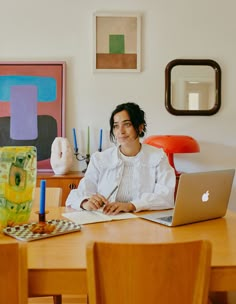 a woman sitting at a table with a laptop computer in front of her and an art work on the wall behind her