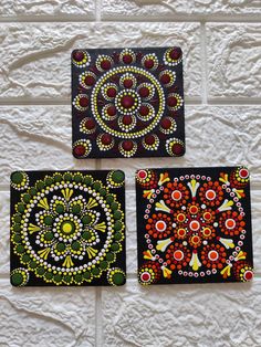 three square paintings are hanging on a white brick wall, one is decorated with beads and the other has bead work