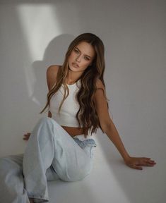 a beautiful young woman sitting on top of a white floor next to a wall with her legs crossed