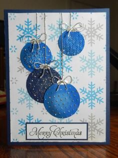 a christmas card with three blue ornaments hanging from it's sides and snowflakes in the background