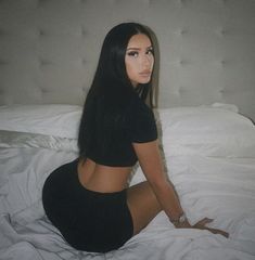 a woman sitting on top of a bed in a black shirt and short skirt next to a white headboard