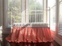 a white crib with pink ruffled bedspread in front of two windows