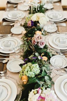 a long table is set with white plates and silverware, flowers and greenery