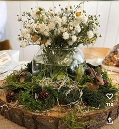 a vase filled with white flowers and greenery on top of a wooden tablecloth