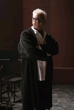 an older man with white hair and glasses is standing in front of a chair, arms crossed