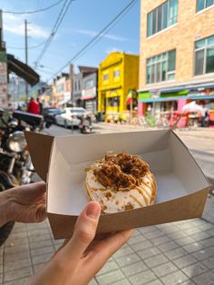 a person holding up a donut with toppings in a box on the street