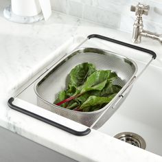 a colander filled with green leafy greens in a kitchen sink next to a faucet