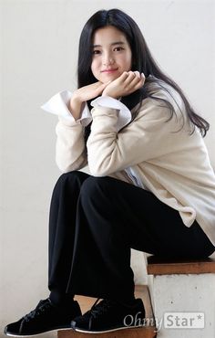 a woman sitting on top of a wooden bench next to a white wall with her hands under her chin