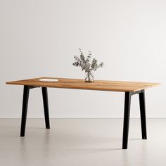 a wooden table with two black legs and a flower vase on the top, in front of a white wall