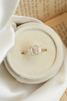 an engagement ring with three baguettes sits on top of a white cloth covered box