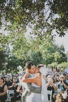 a bride and groom kissing under a tree in front of an outdoor wedding ceremony crowd