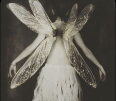 a woman in white dress with wings on her head and body, standing against a dark background