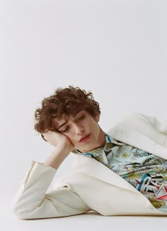 a young man with curly hair wearing a white blazer and colorful shirt sitting on the floor