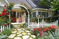 a white picket fence surrounded by flowers and greenery in front of a small house