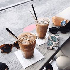 two iced coffees are sitting on a table with sunglasses and cell phone in the background