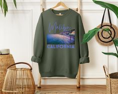 Get ready to ride the waves of style with our Malibu California sweatshirt! This amazing design features a captivating 3D-like graphic of Surfrider Beach at night, capturing the allure of this iconic surf spot. The word "Malibu" is elegantly written above in a playful and elegant script font, while "California" boldly stands below in a bold and stylish san-serif font, both in the same eye-catching color. Whether you're a beach lover, a surf enthusiast, or simply captivated by the coastal charm of Malibu, this sweatshirt is a must-have addition to your wardrobe. So, catch the cool California vibes and rock this stylish and cozy sweatshirt wherever your Malibu adventures take you! Designed with a perfect blend of soft cotton and breathable material, this classic heavy-blend crewneck sweatshi Casual Winter Top With Custom Print, Casual Winter Sweatshirt With Front Print, Casual Sweatshirt With Front Print And Relaxed Fit, Casual Cotton Sweatshirt, Casual Streetwear Sweatshirt For Beach Season, Relaxed Fit Sweatshirt For Beach Season Streetwear, Casual Cotton Sweatshirt For Surfing, Graphic Print Crew Top For Surfing, Casual Green Sweatshirt With Custom Print