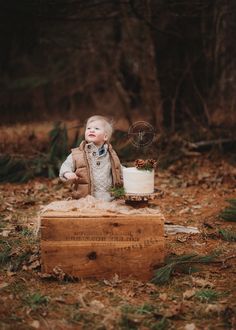 a baby boy sitting on top of a wooden box in the woods with a cake