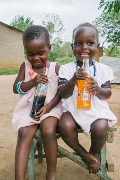 two young children sitting on a bench with bottles in their hands and smiling at the camera