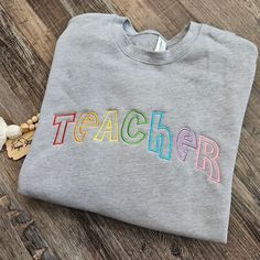 Welcome to my shop! This listing is for an embroidered crewneck sweatshirt!  *Sweatshirt is a Bella Canvas drop shoulder pullover in heather gray. Sizing runs unisex, these are a high quality sweatshirt with long sleeves and torso (see size chart in photos). Other sweatshirt colors may be done upon request for an additional fee. *Front design is TEACHER in rainbow colors!  Feel free to message me for any custom requests! Can change any wording or colors! PaigesStitches Shop Notes: Thank you for Gray Letter Embroidery Sweatshirt For Fall, Gray Cotton Sweatshirt With Letter Embroidery, School Crew Neck Sweatshirt With Embroidered Logo, Customizable Crew Neck Sweatshirt For School, Crew Neck Sweatshirt With Embroidered Graphics For School, Graphic Embroidered Crew Neck Sweatshirt For School, Embroidered Crew Neck Sweatshirt For School, Embroidered Gray Crew Sweatshirt, Gray Cotton Sweatshirt With Embroidered Graphics