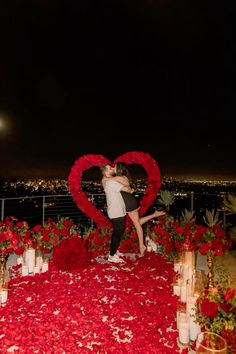 a couple kissing in front of a heart - shaped arrangement of red flowers and candles