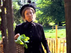 an older woman in a black dress and hat is holding some green leafy plants