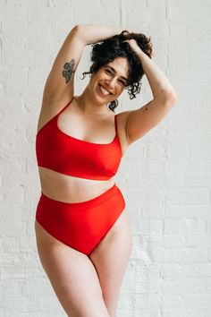 A woman leaning to the side with her hands in her hair wearing bright red high waisted bikini bottoms with full coverage and a matching bright red bikini top. Red High Stretch Sports Bra With Built-in Support, Red Sports Bra With Seamless Stretch Construction, Fitted Sports Bra With Scoop Back For Summer, Red Seamless Stretch Sports Bra, Seamless High Stretch Scoop Neck Swimwear, Red Fitted Sports Bra For Summer, Fitted Red Sports Bra For Summer, Red Stretch Scoop Neck Swimwear, Stretch Swimwear With Bra Friendly Wide Straps