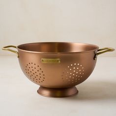 a large metal bowl with handles and holes on the side, sitting on a white surface