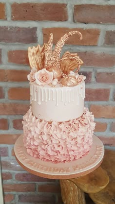 a three tiered cake with pink flowers and gold leaves on top, next to a brick wall