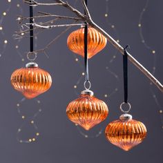 three orange glass ornaments hanging from a branch with lights in the back ground behind them