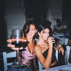 two women sitting at a table with wine glasses in front of them and people standing around