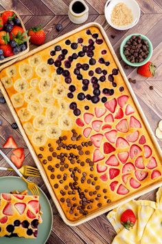 a pancake with fruit and chocolate chips on it next to bowls of berries, bananas, strawberries