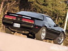 the rear end of a black sports car parked in front of some trees and bushes