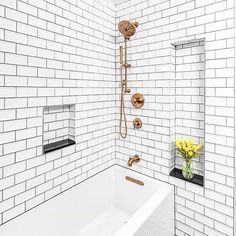 a white tiled bathroom with a tub, shower head and yellow flowers in the vase