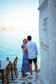 a man and woman standing next to each other near the ocean with their arms around each other