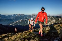 two men are running up a hill on a sunny day with mountains in the background