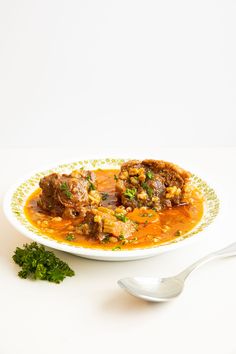 a white plate topped with meat and tomato soup next to a small green leafy garnish