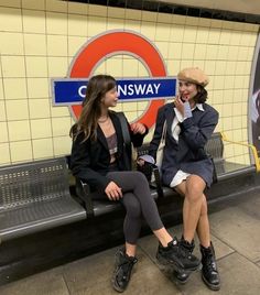 two women sitting on a bench in front of a subway station sign talking to each other