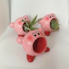 three pink plastic toy pigs with plants in their mouths on a white surface, one has its mouth open and the other is sticking out