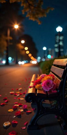 a wooden bench with flowers on the ground next to it and lights in the background