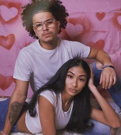 a man and woman sitting on top of a bed in front of a pink wall