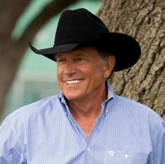 an older man wearing a cowboy hat standing next to a tree smiling at the camera