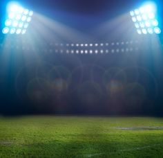 an empty soccer stadium with bright lights and grass in the foreground is lit by floodlights