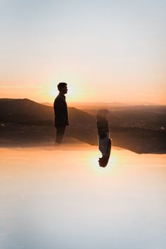 two people standing on the edge of a body of water at sunset, with their reflection in the water