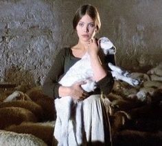 a woman holding a baby in her arms while standing next to some sheep on the ground