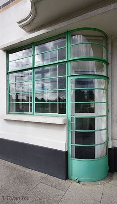 a green window on the side of a white building next to a sidewalk with a red fire hydrant in front of it