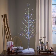 a small christmas tree with white lights and presents on a table in front of a window