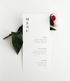 a white menu card sitting on top of a table next to a pink flower and green leaves