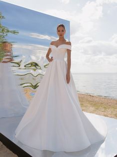 a woman standing in front of a painting wearing a white wedding dress with an off the shoulder neckline