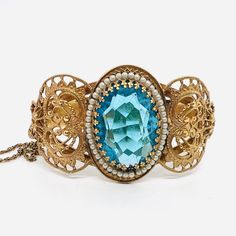 Gorgeous Vintage/Antique Wide Ornate Victorian Revival Blue Topaz Glass Bangle Bracelet. Gold tone/plated ornate filigree metal set with large topaz glass stone surrounded by faux seed pearls. In excellent vintage condition with minimal age appropriate wear. Measures 1 3/8 inches wide in from and 1/2 inch wide in back. Opening is 2 1/4 inches across. Has secure push in clasp and it's original safety chain Blue Intricate Design Jewelry Bracelet, Metal Filigree Bangle Jewelry, Victorian Topaz Jewelry For Formal Occasions, Ornate Adjustable Jeweled Jewelry, Ornate Metal Bracelets With Jewels, Antique Jeweled Bangle Jewelry, Antique Jeweled Bangle, Antique Topaz Jewelry, Victorian Gold Jeweled Bracelets